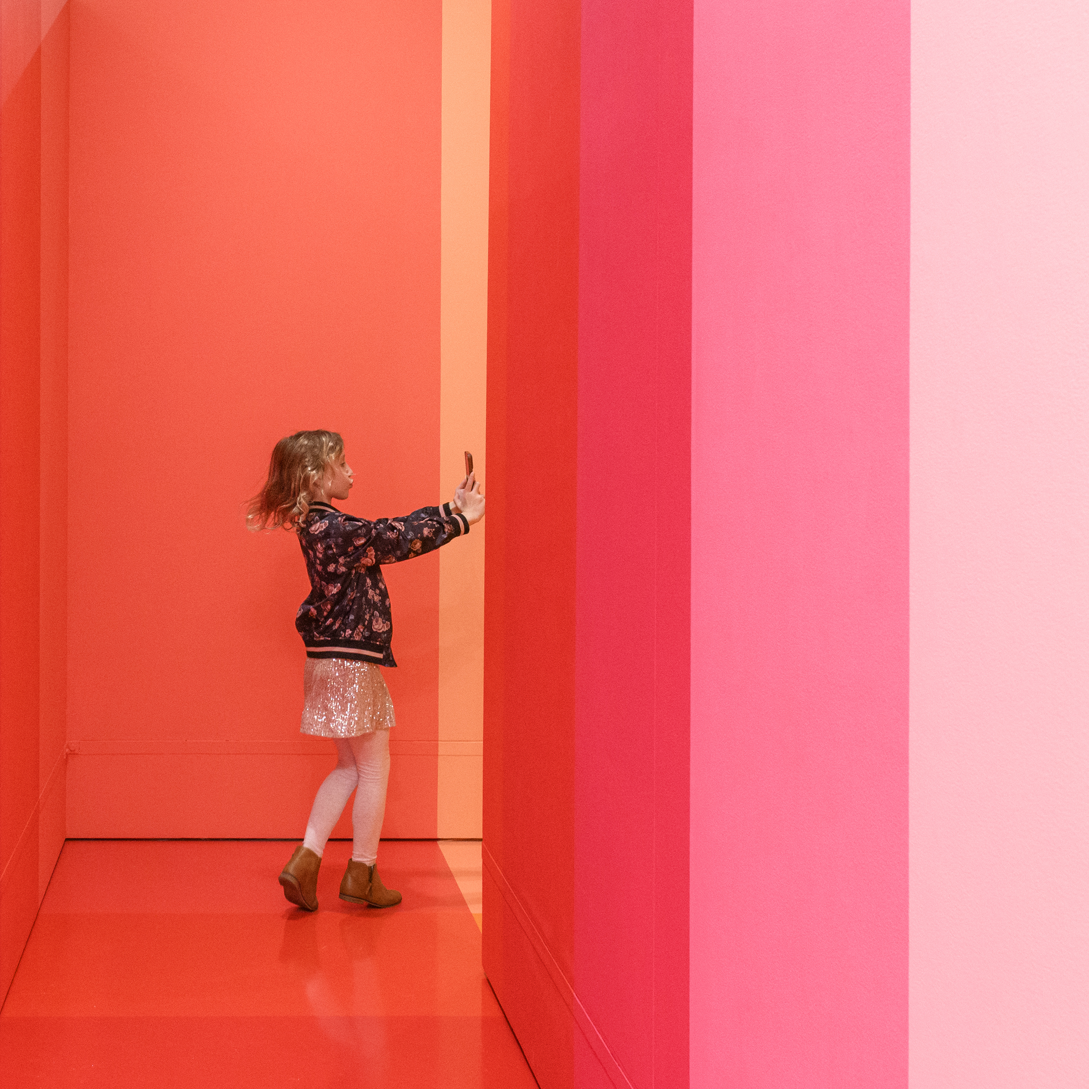 A young girl stands in a vibrant, pink and coral-colored hallway, reaching up towards the wall. She is wearing a floral jacket, a white skirt, and brown boots. The minimalist setting with its intense vertical stripes exudes the bold, colorful ambiance of a Denver Design Studio creation.