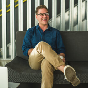 A smiling man with glasses, in a blue shirt and beige pants, sits on a dark gray sofa with one leg crossed over the other. He is holding a cup and appears to be laughing. Behind him are the sleek metal stairs with a yellow handrail crafted by Denver Design Studio.