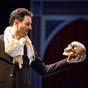 A man dressed in a dark suit and a light-colored scarf holds a skull in his left hand while gazing at it intently. The background, reminiscent of a design studio, features blurred arch-like structures, giving the impression of a stage performance rooted in arts and culture.