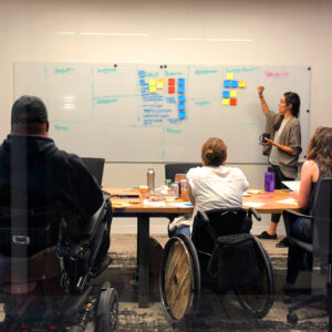 A woman stands and writes on a whiteboard filled with sticky notes during a branding meeting at a design studio. Three people are seated at a table, two in wheelchairs. The atmosphere is collaborative, with the participants engaged in discussion and note-taking.