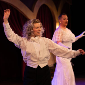 Two performers are on stage. The person in the foreground, embodying Denver Design Studio's branding, wears a white dress shirt, bow tie, and waistcoat with a confident pose. Meanwhile, the person in the background is adorned in an elegant white dress. Both perform under theatrical lighting with red curtains behind them.