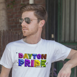 Person with short brown hair and wearing sunglasses is leaning against a railing. They are wearing a white t-shirt with the text "DAYTON PRIDE 2024" in colorful, block letters. The background includes a tree and building entrance.