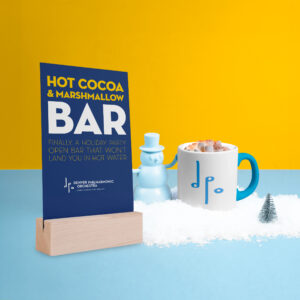 A sign reads "Hot Cocoa & Marshmallow Bar: Finally, a holiday party open bar that won't land you in hot water," by the Denver Philharmonic Orchestra. It's displayed next to a mug of hot cocoa topped with marshmallows, a toy snowman, and snow on a blue and yellow surface.