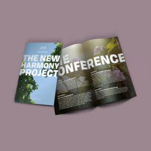 A brochure with the title "The New Harmony Project" on the cover, featuring foliage against a clear sky. The brochure is open to a page with the heading "Conference" and several text sections describing different programs. The background is a solid muted purple.