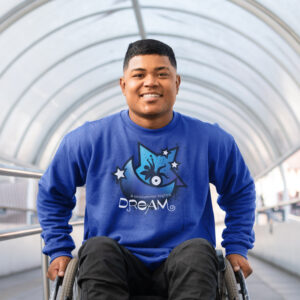 A young man in a wheelchair smiles while wearing a blue sweatshirt with a design referencing "A Midsummer Night's Dream." He is positioned in a bright, curved corridor.