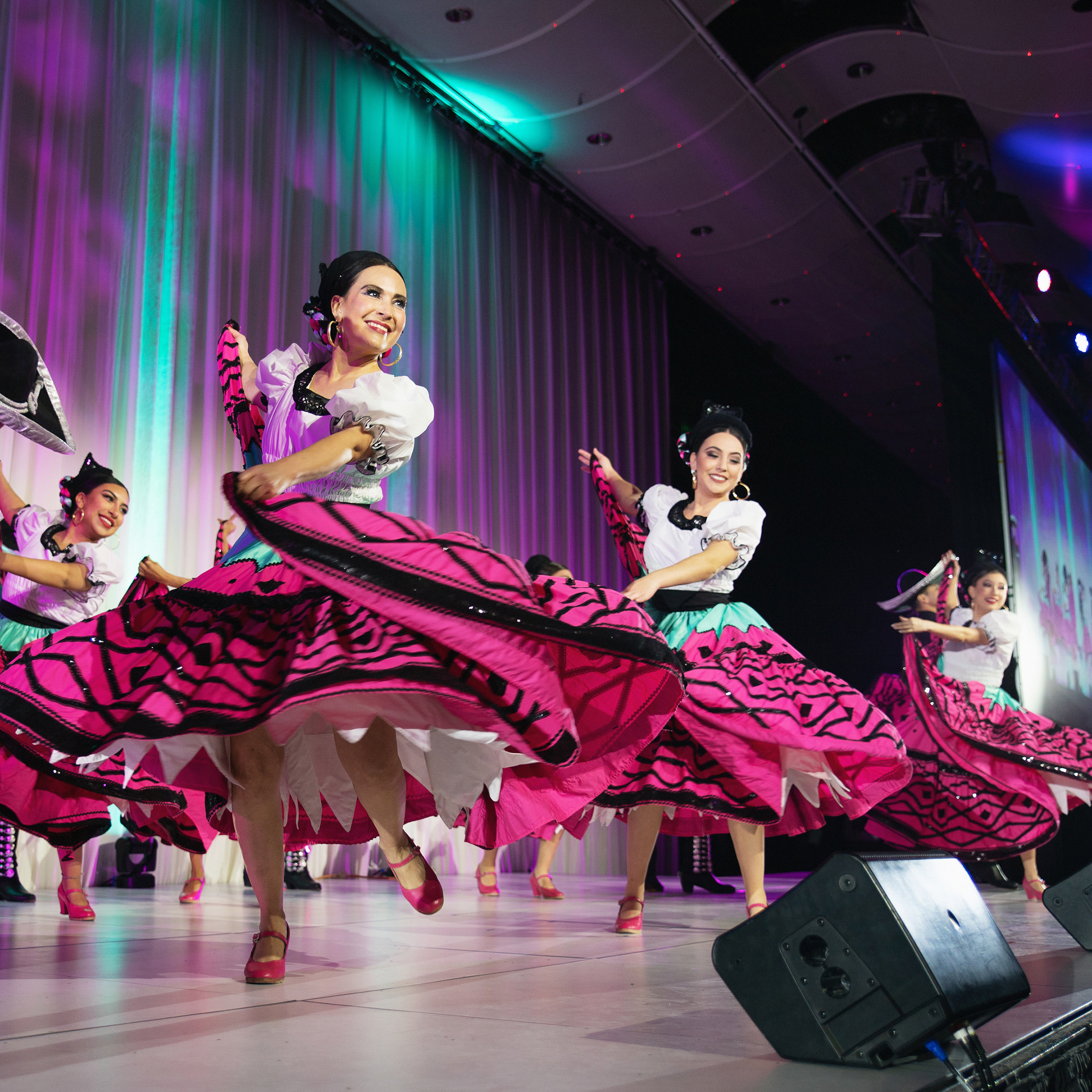 A group of dancers in colorful traditional outfits perform on stage with bright lighting.