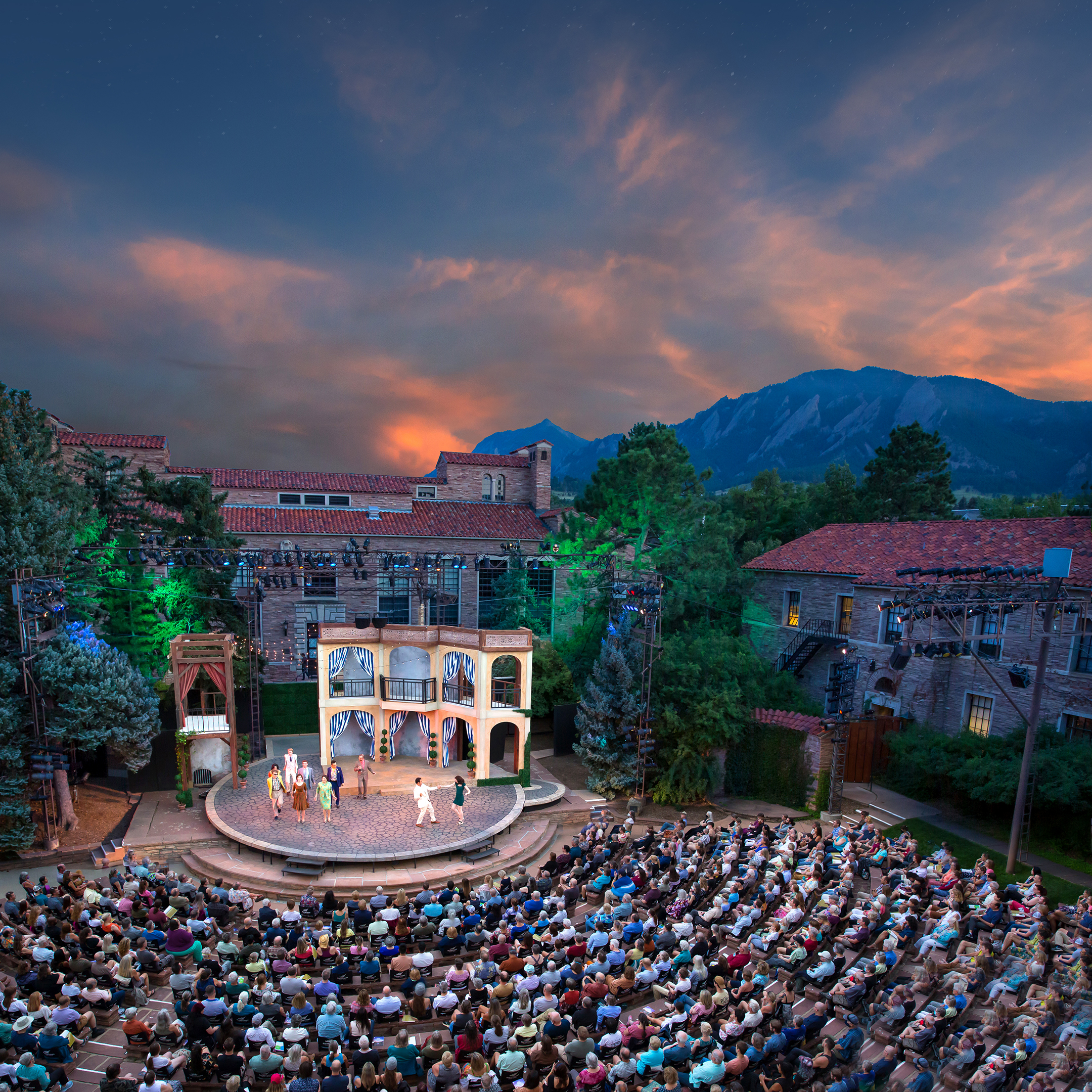 Outdoor theater performance at dusk with an audience seated in a semicircular arrangement. The stage features a multi-level set, with mountains and a colorful sky in the background.