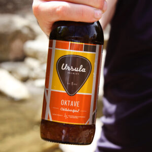 Close-up of a hand holding a bottle of Ursula Brewery Oktave Oktoberfest beer. The label displays orange, yellow, and brown colors with geometric patterns.