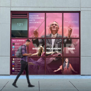 A man walks past a window display for Dayton Performing Arts Alliance, featuring a conductor, ballet dancer, and soprano. The display highlights the Philharmonic's 30th season.