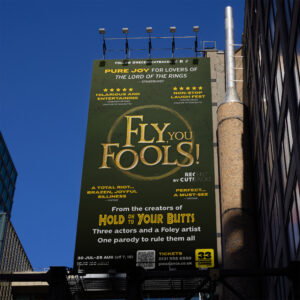 A large outdoor billboard advertises a parody show called "Fly You Fools!" with comedic quotes and show details against a green background, mounted on the side of a building.
