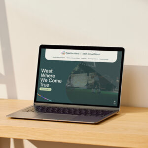 A laptop on a wooden surface displays the Creative West 2024 Annual Report website, with the text "West Where We Come True" visible on the screen.