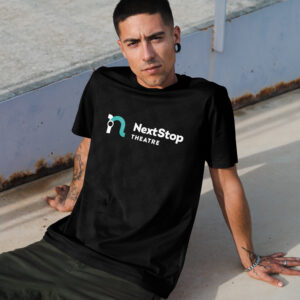 A man with tattoos and stretched earlobes sits on the ground, wearing a black "NextStop Theatre" t-shirt.