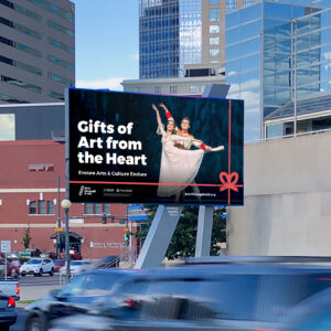 A digital billboard in a city displays an ad featuring two ballet dancers and the message: "Gifts of Art from the Heart. Ensure Arts & Culture Endure.