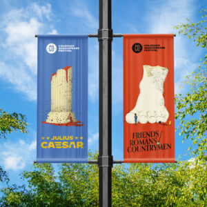 Two vertical festival banners on a pole; one features a broken Roman column and "Julius Caesar," the other a marble foot and "Friends Romans Countrymen," both with Colorado Shakespeare Festival logos.