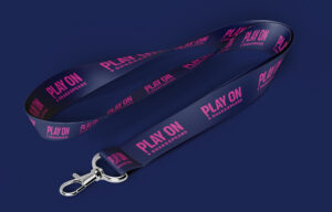 A navy blue lanyard with "PLAY ON SHAKESPEARE" printed in bold pink letters and a silver clasp, displayed on a solid dark blue background.